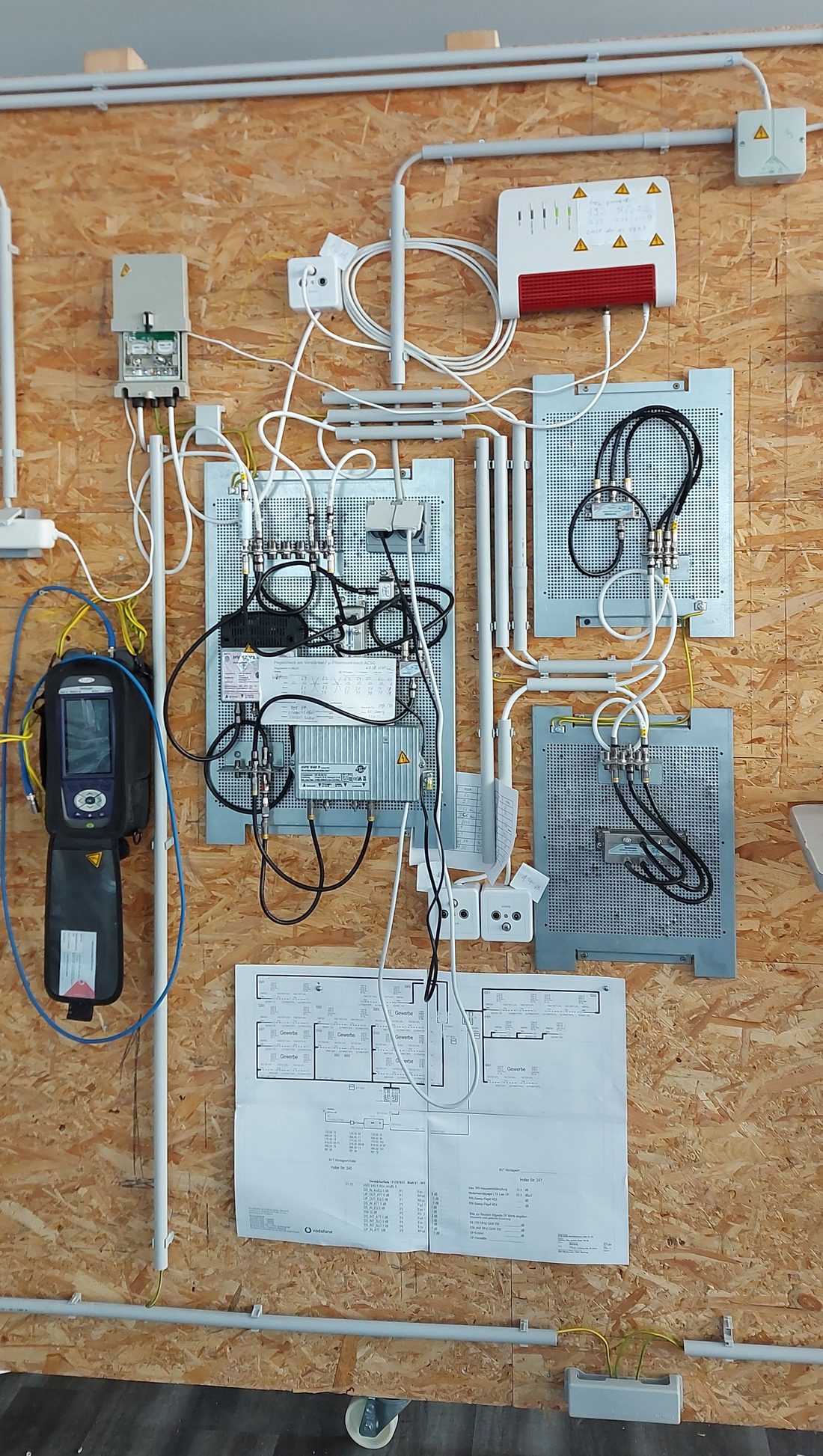 Koax-Schulungsanlage an einer Holzwand mit Hausübergabepunkt, Verstärker, Verteilern, Anschlussdosen, Kabelrouter und Messgerät